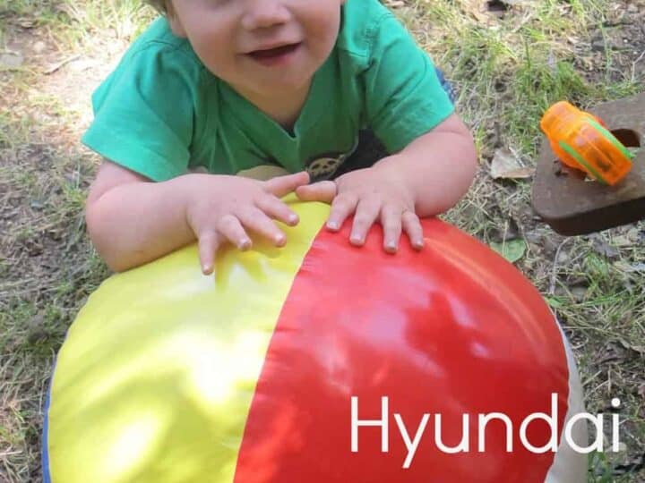A toddler in a green shirt beams with joy while playing with a large, colorful beach ball outdoors at the Hyundai Epic Playdate. Vibrant text highlights the event's playful spirit.