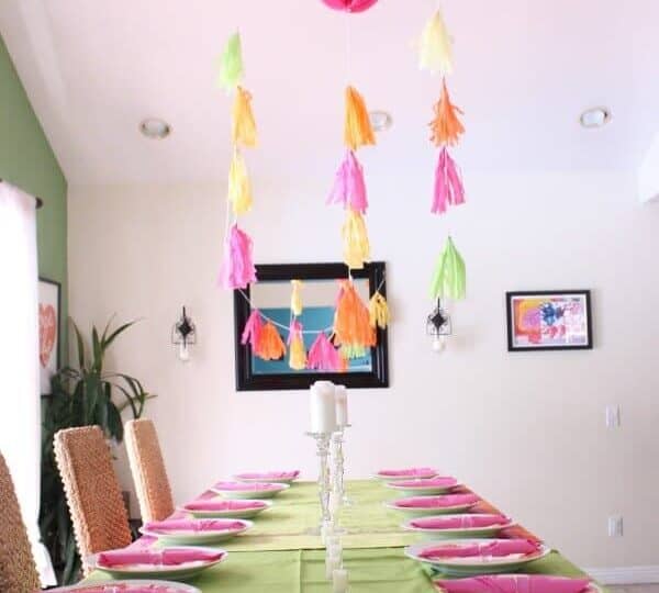 A brightly decorated dining table set for a baby shower, featuring a green tablecloth and pink napkins on white plates. Colorful paper decorations and tassels hang above, adding to the vibrant atmosphere, with framed artworks adorning the cream-colored walls.