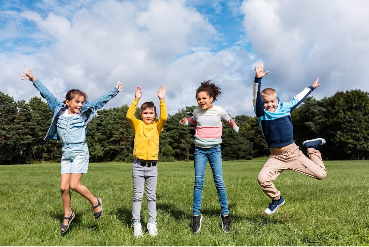 Four children are jumping and smiling in a grassy field with trees in the background. The sky is partly cloudy, and the kids look happy and energetic, wearing casual clothing in bright colors.