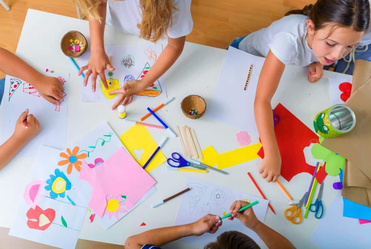 Four children sitting around a table are creating colorful crafts with paper, glue, markers, scissors, and other art supplies. The table is covered with drawings, cutouts, and various craft materials.