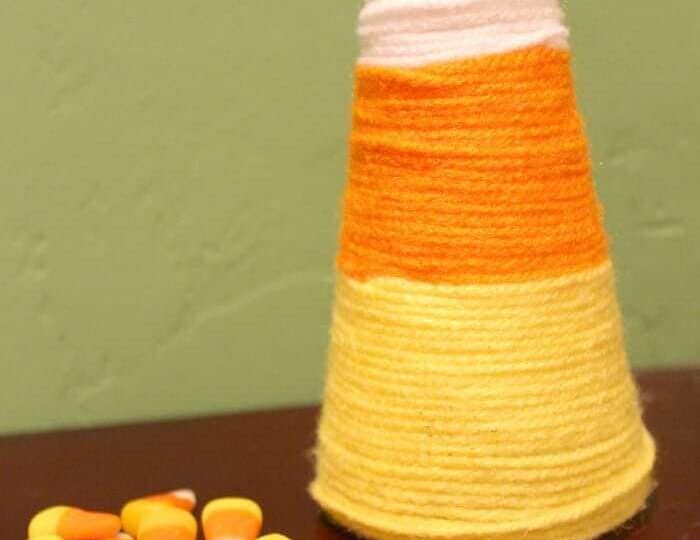 On a table, a Candy Corn Yarn Tree displays its distinctive white, orange, and yellow sections. Scattered nearby are real candy corn pieces, echoing the tree's festive autumn charm.