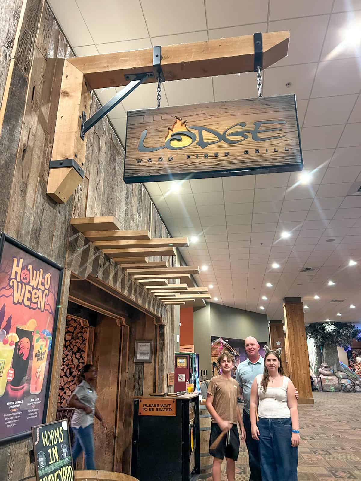 Family standing under a wooden sign for Lodge Wood Fired Grill in a rustic-themed indoor area of the Great Wolf Lodge in Southern California