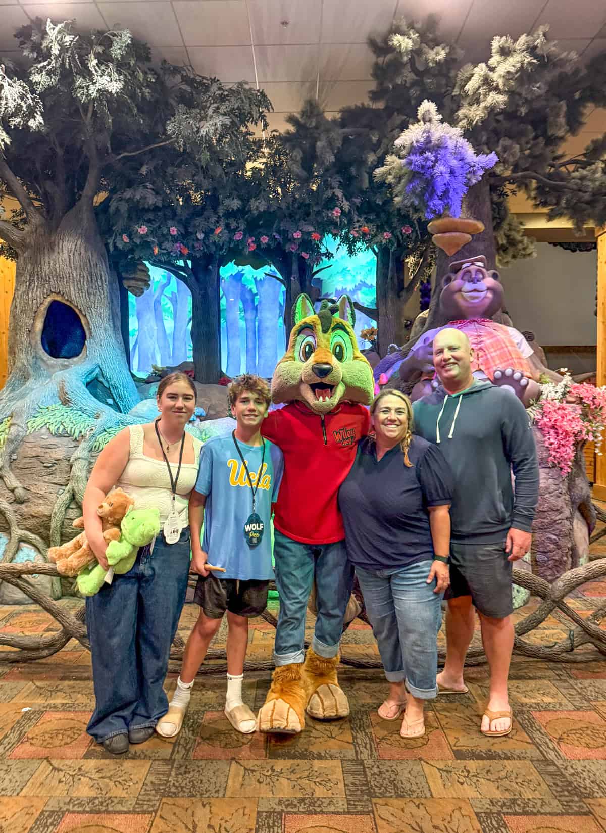 Family pose with wolf Character in front of a forest-themed backdrop at Great Wolf Lodge Southern California. Everyone is smiling and standing close together&mdash;proof that Is Great Wolf Lodge Fun for Teens? Absolutely!.