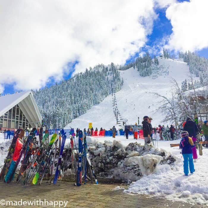 At Squaw Valley, a snowy ski resort, people in colorful gear gather at the base of the mountain. Skis and poles are lined up in the foreground as a ski lift ascends amid snow-covered trees, all under a bright blue sky with scattered clouds.