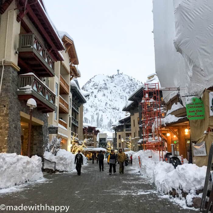 A snowy street in Squaw Valley features people strolling between charming buildings. Snow-covered mountains rise majestically in the background, while scaffolding adorns one of the structures, adding a touch of rustic development to the wintry, picturesque scene.