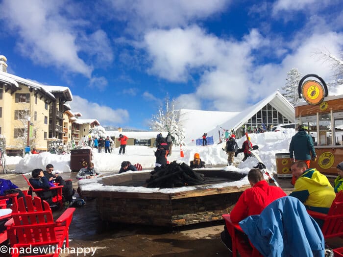 People gathered around an outdoor fire pit near the snowy ski resort at Squaw Valley. Red chairs surround the fire pit, and skiers are seen walking in the background under a bright blue sky.