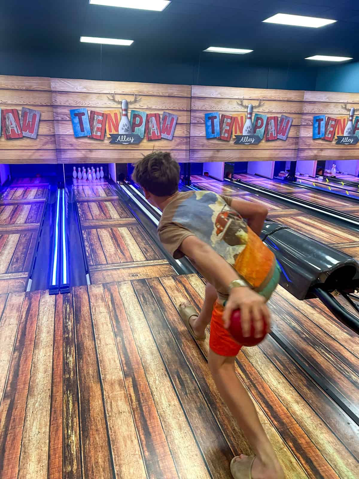 A teen wearing a colorful T-shirt and orange shorts bowls a red ball down a wooden lane at Ten Paw Alley, an indoor bowling alley with bright lights and vibrant décor.