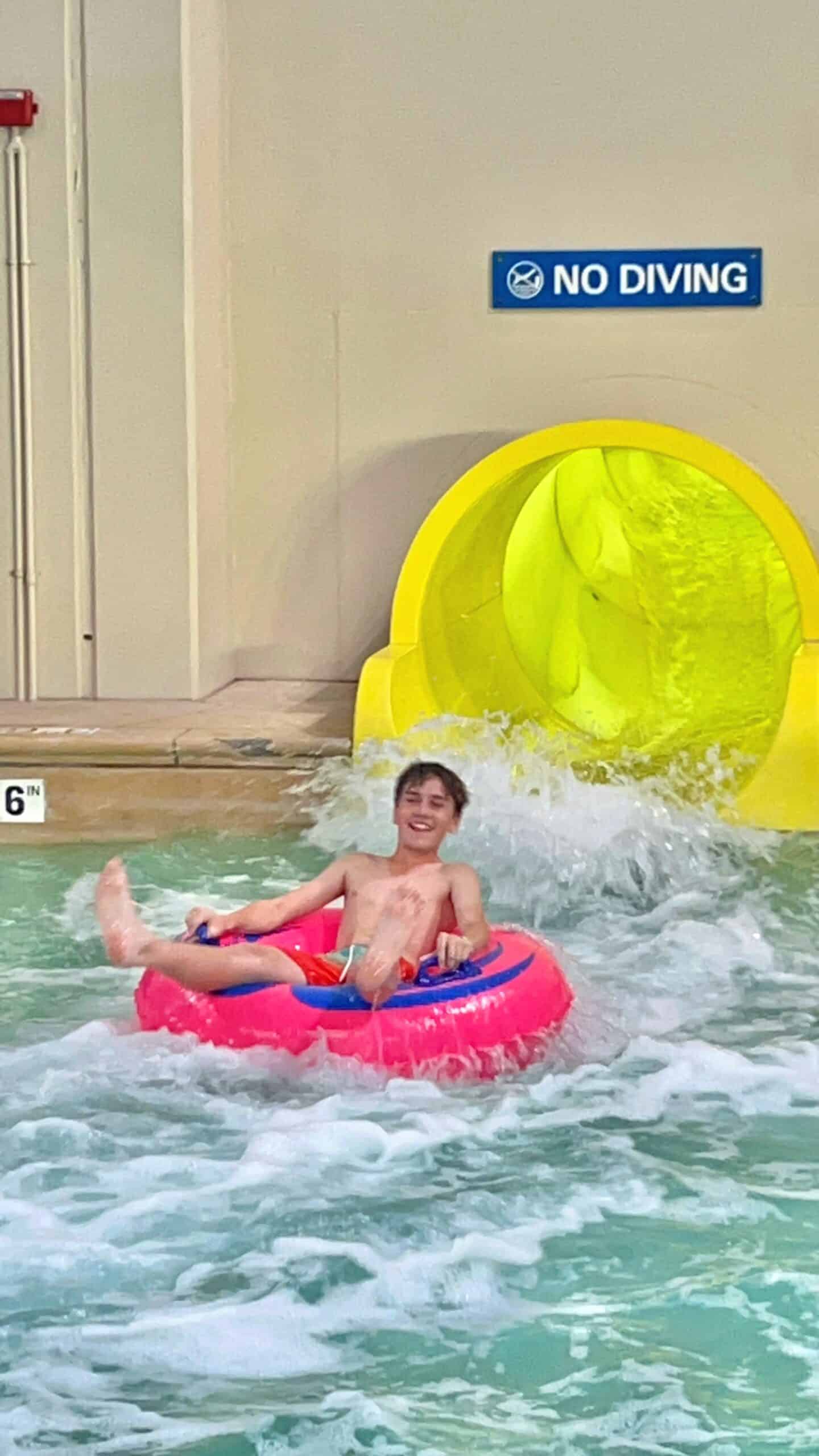 A smiling teen boy in swim trunks rides a pink inner tube in a pool, exiting a yellow water slide. Water splashes around him and a No Diving sign is visible on the wall above the slide.