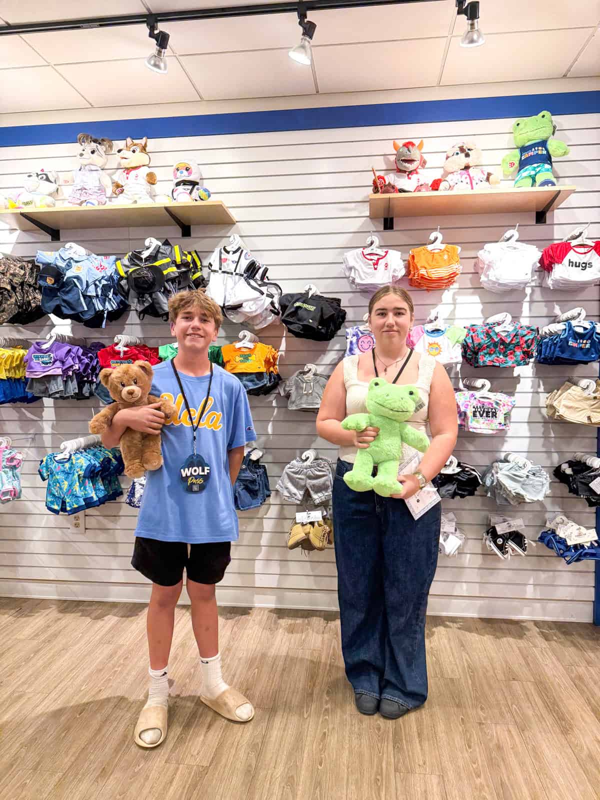 Teens stand inside a Build-a-Bear store within the Great Wolf Lodge Southern California holding stuffed animals. The person on the left holds a brown teddy bear and wears a blue shirt, shorts, and slippers. The person on the right holds a green stuffed animal. Plush toys and outfits are displayed behind them.