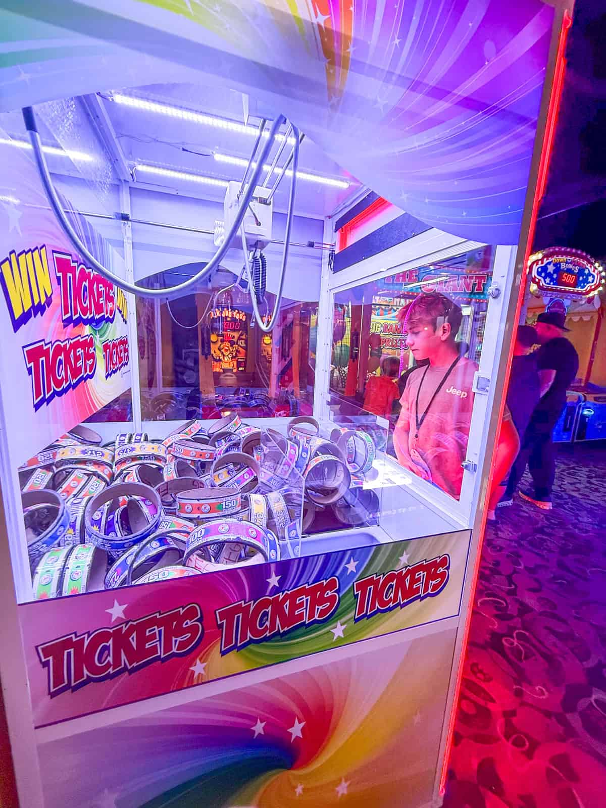 A teen stands in front of a brightly lit arcade claw machine filled with rolls of tickets, surrounded by colorful lights and arcade games at The Great Wolf Lodge Southern California