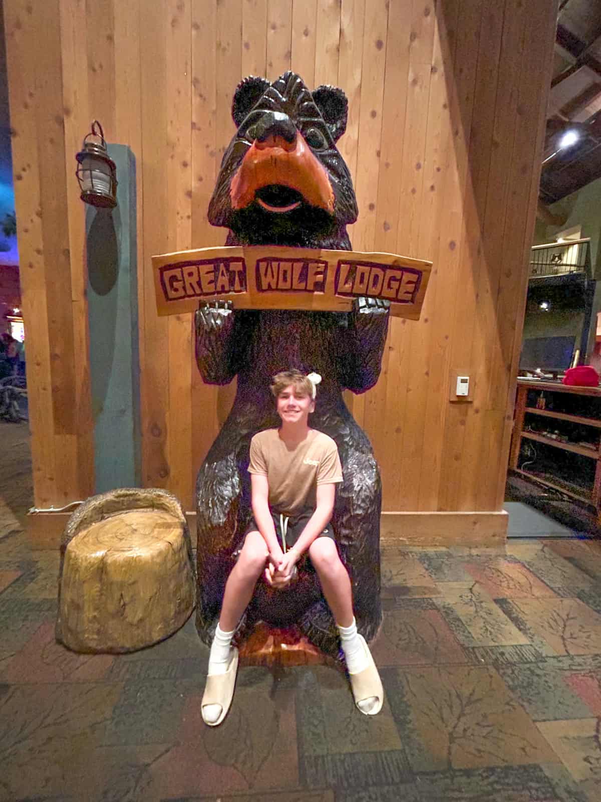 A smiling teens sits on the lap of a large black bear statue holding a Great Wolf Lodge sign inside a rustic, wood-paneled building.