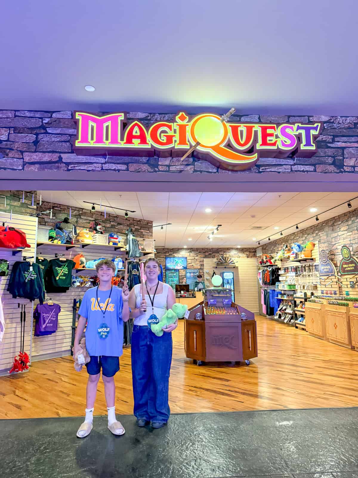 Two teenagers stand smiling under a colorful “MagiQuest” sign at an indoor shop, holding stuffed animals and wearing medals. Shelves behind them display clothing and various merchandise.