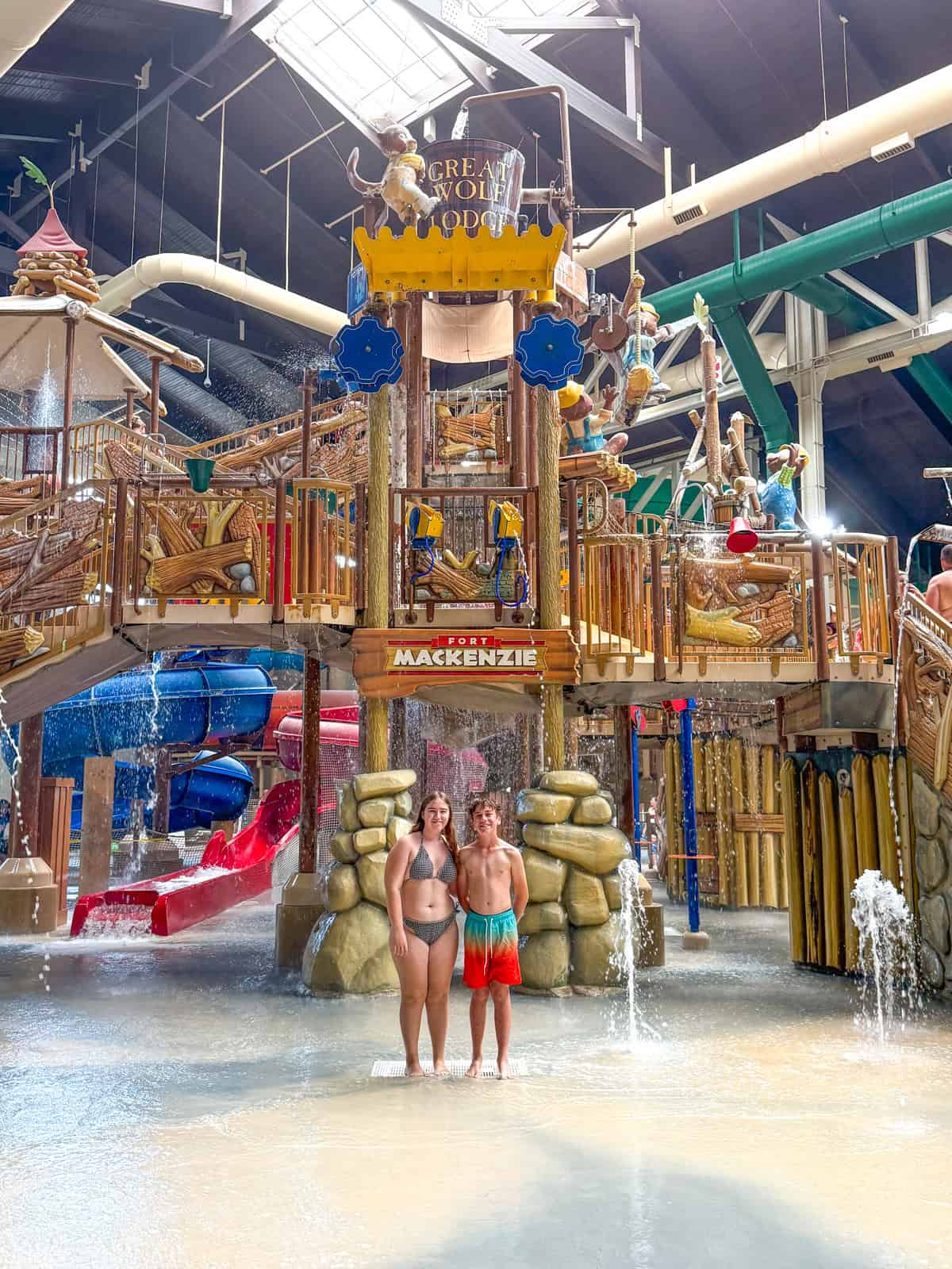 Teens in swimsuits stand in shallow water in front of a large indoor water play structure with slides, stairs, and water features at Great Wolf Lodge Southern California. The area is bright and busy with playful elements all around.