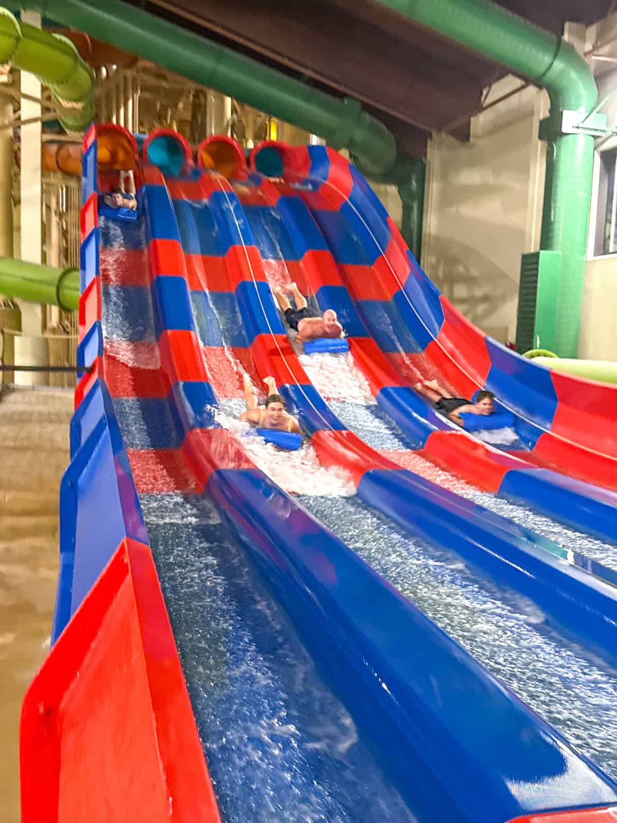 Teens race down parallel red and blue water slides inside an indoor water park, splashing water as they slide on their backs, with green pipes and a high ceiling in the background.