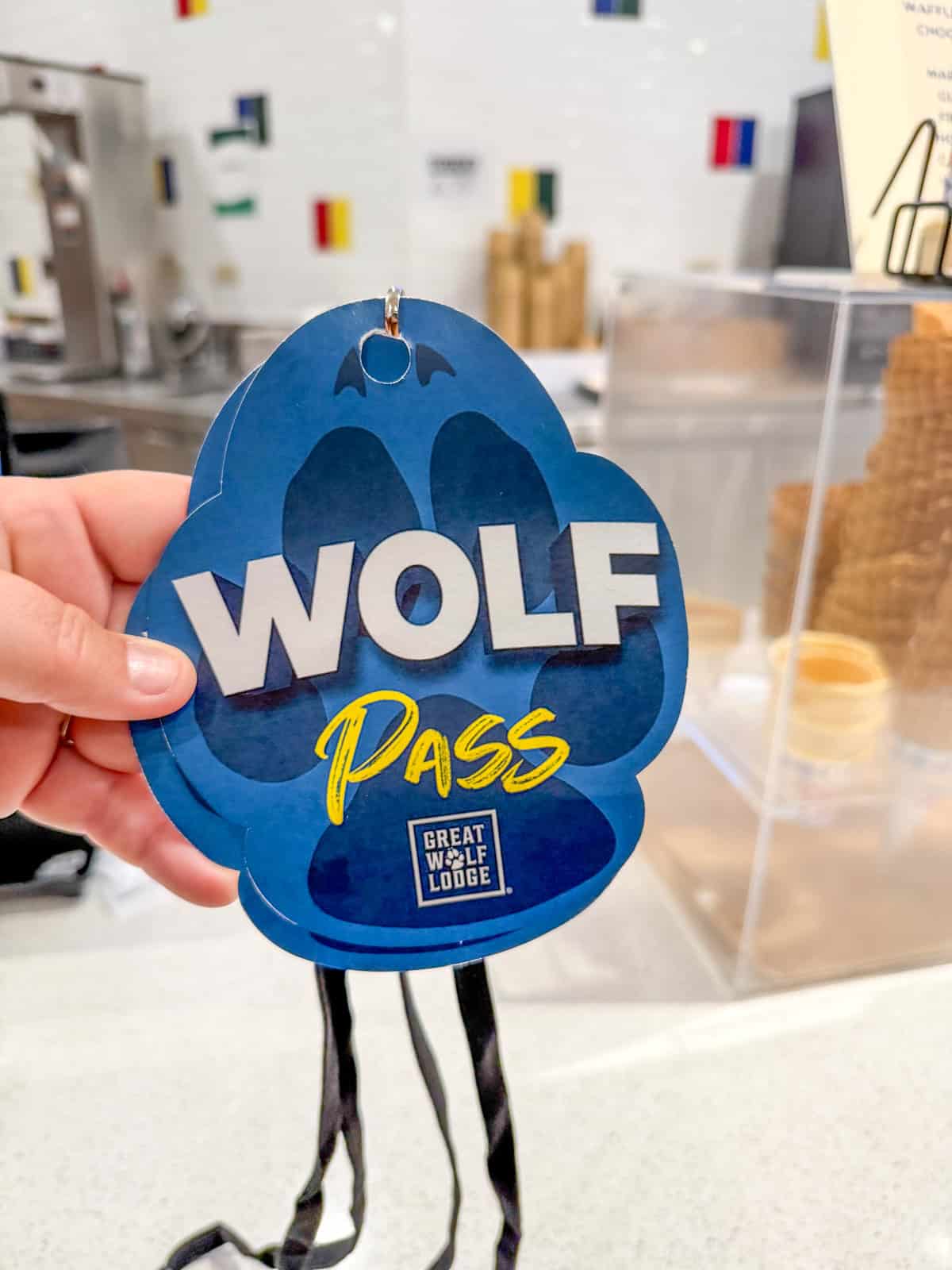 A hand holds a blue, paw-shaped card labeled WOLF PASS from Great Wolf Lodge Southern California, with a background featuring stacked waffle cones and a counter.