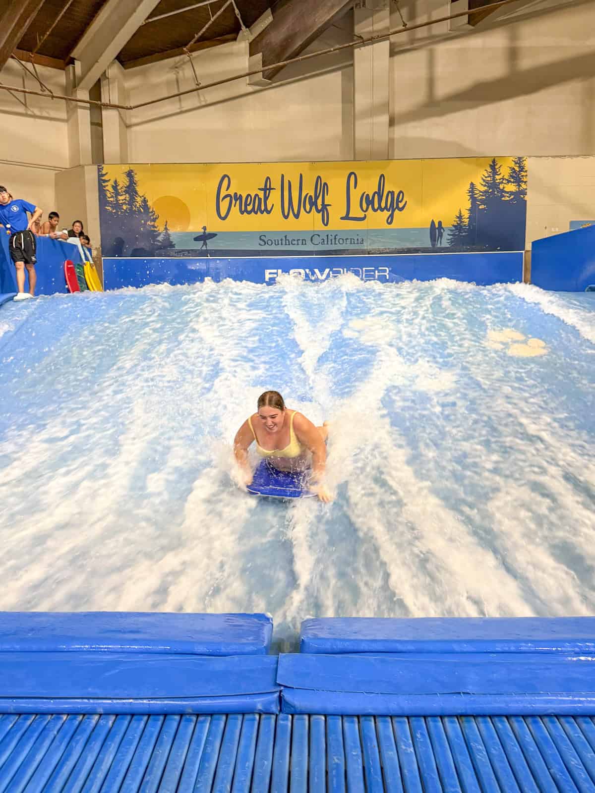 A teen rides a wave on an indoor surf simulator at Great Wolf Lodge Southern California, surrounded by flowing water and splashes under a large blue and yellow sign.