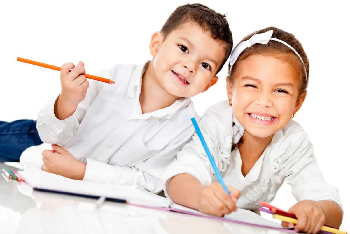 Two children lie on the floor, happily drawing. The boy on the left holds an orange pencil, and the girl on the right holds a blue pencil. Both wear white shirts and smile brightly. Papers and colored pencils are scattered around them.