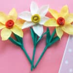 Three handmade paper flowers with yellow and white petals and green stems are arranged on a pink surface with a partially visible white polka dot pattern on the right side.