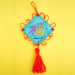 This Good Luck Ornament showcases a decorative Chinese knot set against a yellow background, adorned with the pink character 福 (fu) for good luck, and the words GOOD LUCK beneath. Elegant red tassels gracefully hang from the bottom.