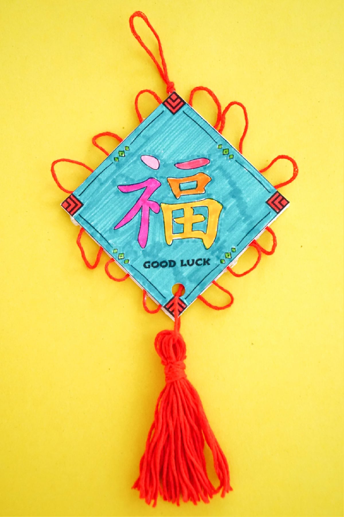 A colorful handmade decoration on a yellow background showcases a blue diamond-shaped card with a red Chinese character and the words GOOD LUCK. This elegant piece of Good Luck Ornaments is adorned with red yarn loops and features a tassel at the bottom.