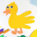 A duck-shaped craft made from yellow paper, with googly eyes and orange beak and feet, is surrounded by markers and paper cutouts on a white background. Blue clouds and leaves are visible nearby.