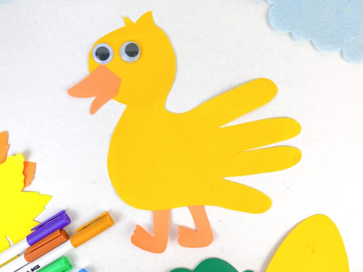 A duck-shaped craft made from yellow paper, with googly eyes and orange beak and feet, is surrounded by markers and paper cutouts on a white background. Blue clouds and leaves are visible nearby.