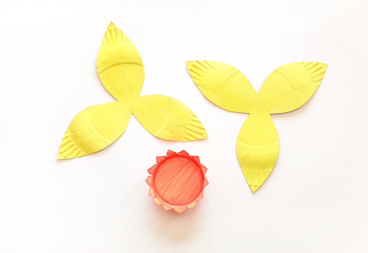 Two yellow paper flower shapes with three petals each are arranged on a white background. Below them is a small red circular object with a zigzag edge, resembling a flower center.