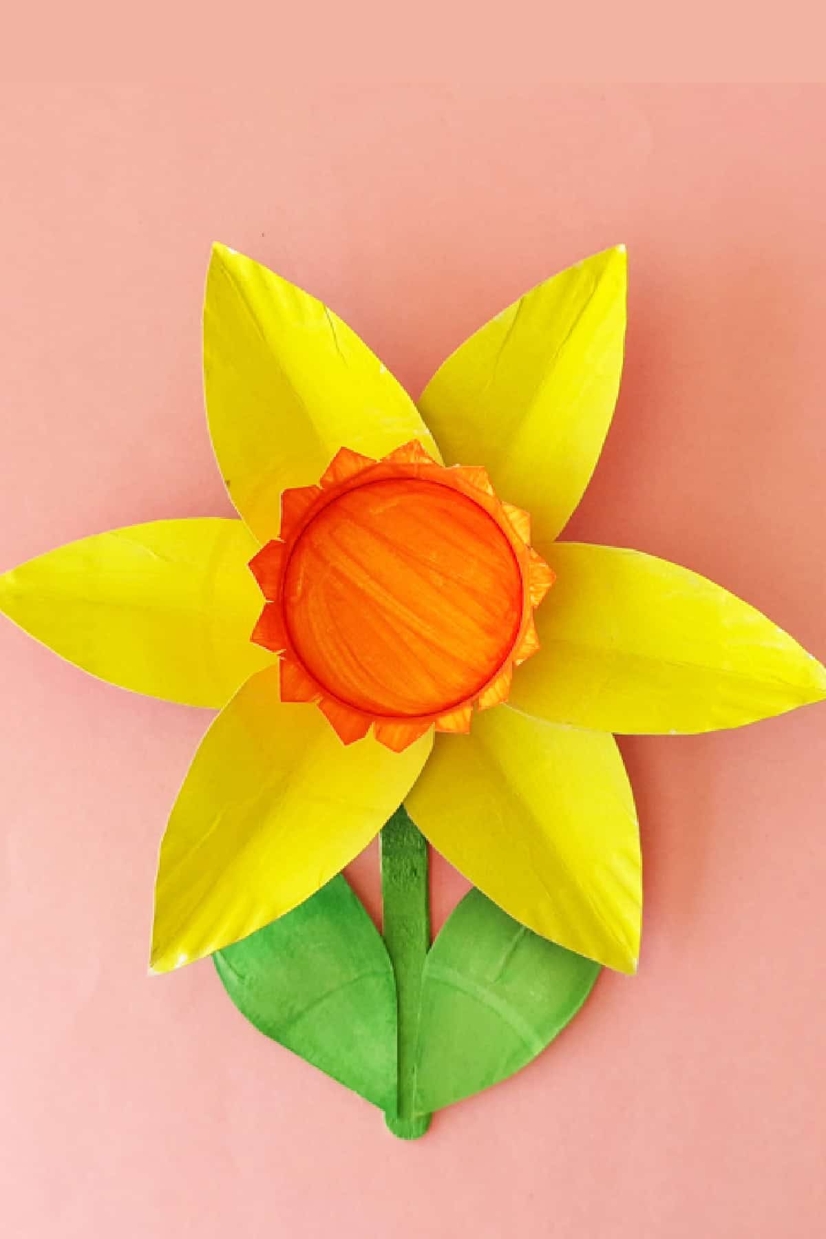 A craft flower made from paper plates on a pink background. The petals are bright yellow, and the center is orange, resembling a daffodil. Green leaves are attached at the bottom.
