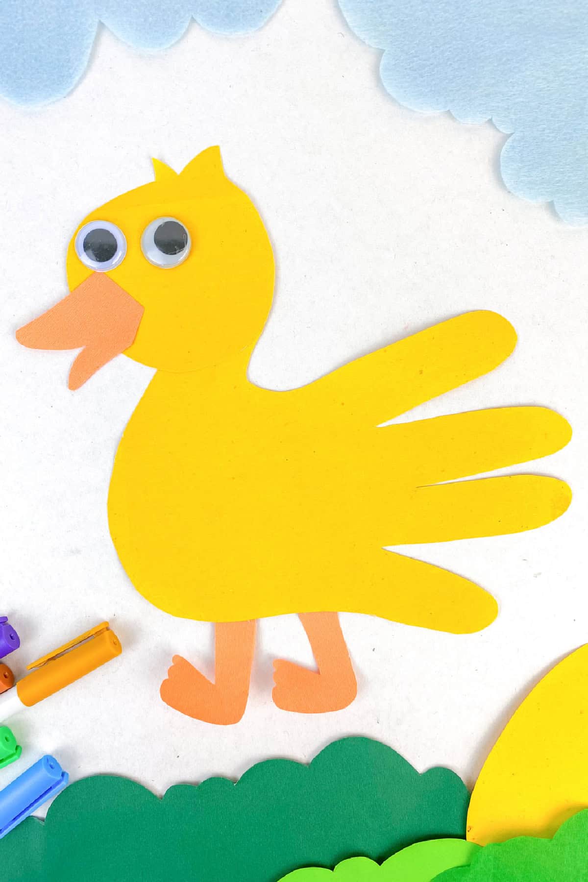 A colorful craft depicting a yellow bird made from paper, using a handprint shape for the body. The bird has googly eyes and an orange beak and feet. Blue clouds and green landscape details are seen around it.