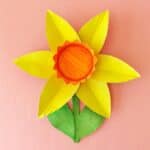 A craft daffodil made from a painted paper plate, featuring bright yellow petals, an orange center, and a green stem with one leaf, set against a pink background.
