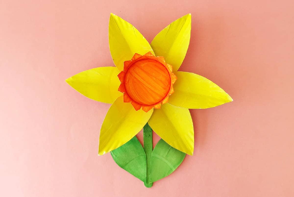 A craft daffodil made from a painted paper plate, featuring bright yellow petals, an orange center, and a green stem with one leaf, set against a pink background.